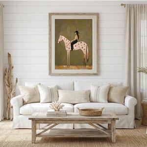 May include: A framed wall art print featuring a stylized illustration of a person wearing a black hat and dark clothing riding a light-colored horse with dark star-shaped markings on its body, set against a muted olive green background.