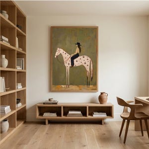 May include: A framed wall art print featuring a stylized illustration of a person wearing a black hat and dark clothing riding a light-colored horse decorated with dark star patterns against a muted olive green background.