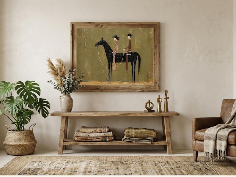 A framed artwork depicts two figures riding a black horse against a green backdrop. The artwork is displayed above a wooden console table, complemented by a potted plant, decorative vase, and a leather armchair, creating a rustic interior design.