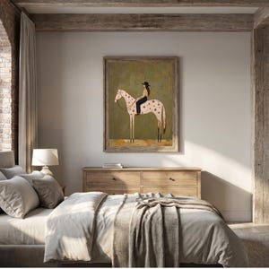 May include: A framed wall art print featuring a stylized illustration of a person wearing a black hat and dark clothing riding a light-colored horse with dark spots. The horse is positioned in profile against a muted olive green background.