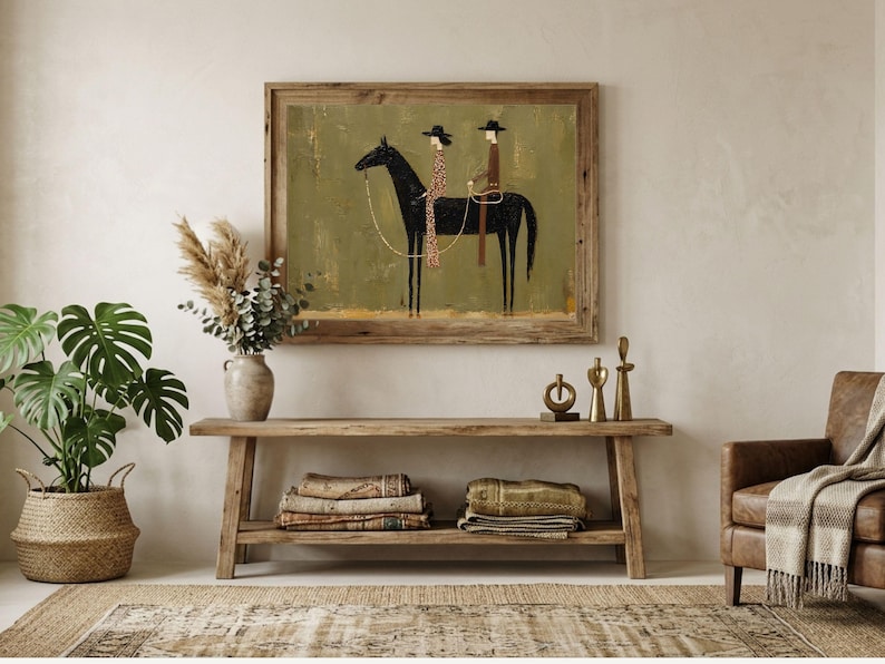 May include: A framed wall art print featuring a stylized illustration of two figures wearing hats riding a black horse against a textured olive green background. The artwork is displayed in a rustic wooden frame, showcasing a minimalist artistic composition.