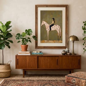 May include: A framed wall art print featuring a stylized illustration of a person wearing a black cowboy hat and dark pants riding a light-colored horse with dark star-shaped markings on its body, set against a muted olive green background.