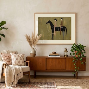 May include: A framed wall art print featuring a stylized illustration of two figures in hats riding a black horse against a muted olive green background. The artwork is displayed in a light wood frame with a wide white mat, showcasing a minimalist aesthetic.