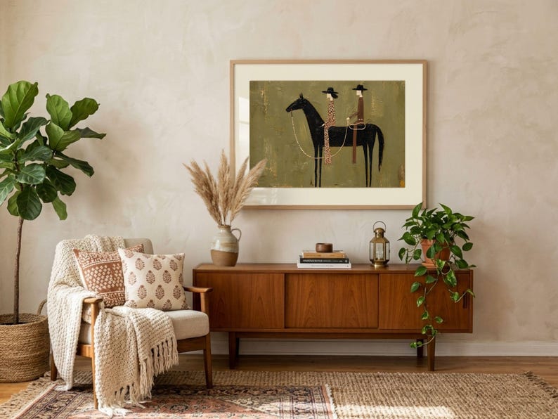 May include: A framed wall art print featuring a stylized illustration of two figures in hats riding a black horse against a muted olive green background. The artwork is displayed in a light wood frame with a wide white mat, showcasing a minimalist aesthetic.