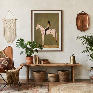 May include: A framed wall art print featuring a stylized illustration of a person in a black hat riding a light pink horse with star patterns against a dark olive green background, displayed in a room with a leather chair and wooden bench.