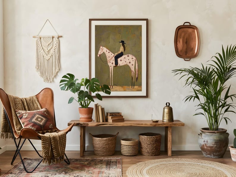 May include: A framed wall art print featuring a stylized illustration of a person in a black hat riding a light pink horse with star patterns against a dark olive green background, displayed in a room with a leather chair and wooden bench.