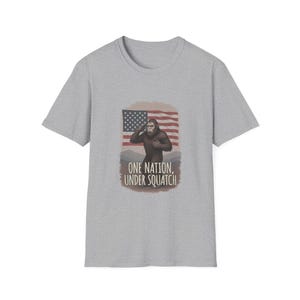 May include: A heather gray short-sleeve t-shirt featuring a graphic of a brown Bigfoot saluting in front of an American flag. Below the figure, the text reads ONE NATION, UNDER SQUATCH in a dark brown, sans-serif font on a light brown background.