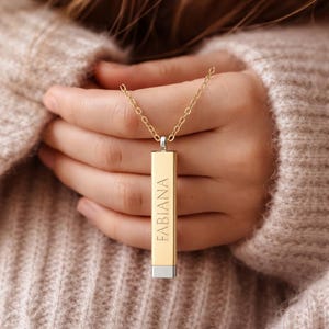 Könnte beinhalten: Eine goldfarbene rechteckige Anhängerhalskette mit dem vertikal eingravierten Namen "FABIANA". Der Anhänger hat einen silberfarbenen Boden und hängt an einer zarten Goldkette. Die Halskette wird vor einem weichen, neutral getönten Hintergrund präsentiert.