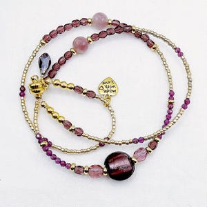 May include: A multi-strand beaded bracelet featuring small gold-tone metallic beads, translucent purple faceted beads, and larger smooth pink and dark purple spherical beads. A small gold-tone heart charm is attached with the text made with love.
