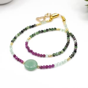 May include: A delicate necklace featuring a large, round, light green stone pendant. The necklace is composed of small, colorful beads in shades of green, purple, and gold, creating a layered effect. The clasp is gold-toned.