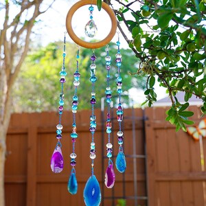 May include: A hanging mobile featuring a circular wooden hoop with a central clear crystal prism. Five strands of blue and purple glass beads hang from the hoop, each ending in a polished, teardrop-shaped agate slice in shades of blue and purple.
