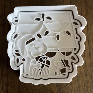 May include: White plastic cookie cutter featuring Snoopy in a Santa hat, holding a present, with Woodstock and Christmas lights. The cutter has a scalloped edge and is designed to cut out a holiday-themed cookie shape.