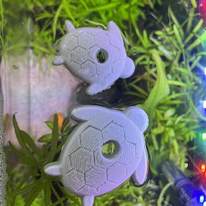 May include: Two light gray turtle-shaped aquarium decorations floating in water. The turtles have a textured shell design with a hole in the center. The background features green aquatic plants and a glimpse of aquarium lighting.