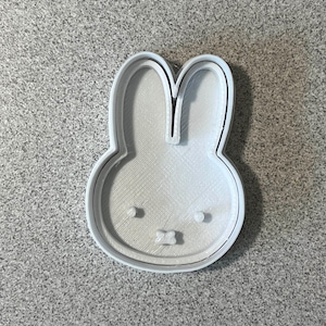 May include: A white plastic cookie cutter in the shape of a bunny head. The cutter has a detailed outline and interior features, including eyes and a bow tie. The design is suitable for making cookies or other treats.