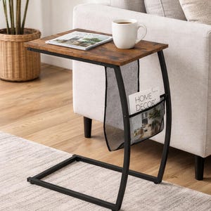 May include: A side table with a dark brown, wood-look top and black metal frame. A white mug with coffee sits on the table, next to a magazine. The table has a mesh pocket for magazines and is positioned next to a light-colored sofa.
