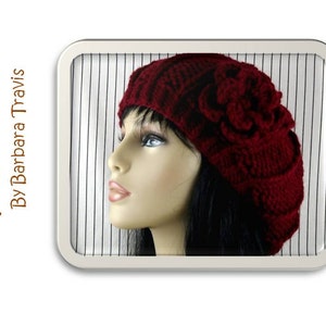 May include: A close-up of a red crocheted beret with a large flower on top. The beret is worn by a mannequin with dark hair.