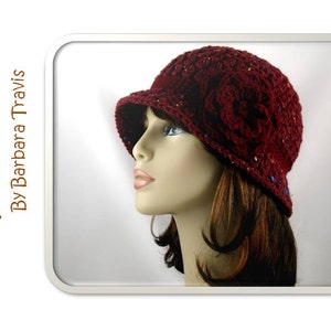 May include: A maroon crocheted hat with a flower embellishment and colorful speckles. The hat is worn by a mannequin with long brown hair.