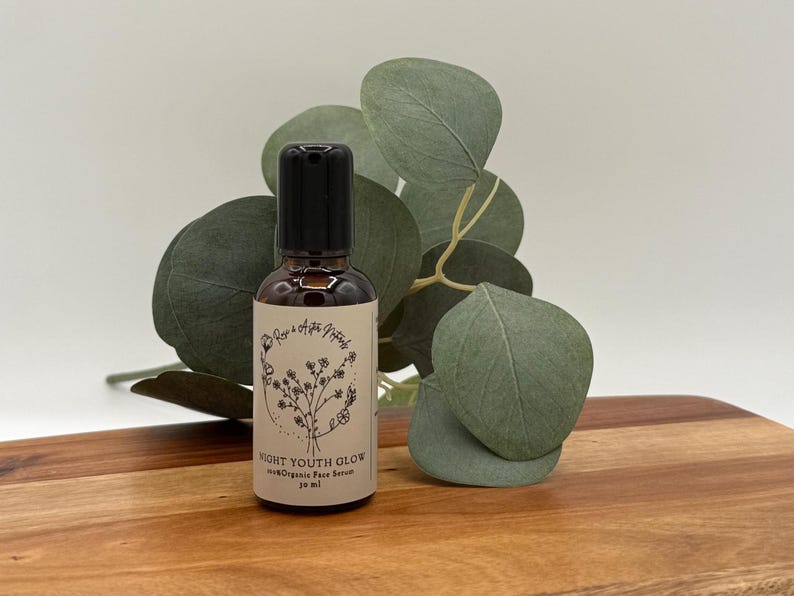 May include: A brown glass bottle with a black cap, labeled "NIGHT YOUTH GLOW 100% Organic Face Serum 30 ml." The bottle is set on a wooden surface, with green leaves in the background. The label has a floral design.