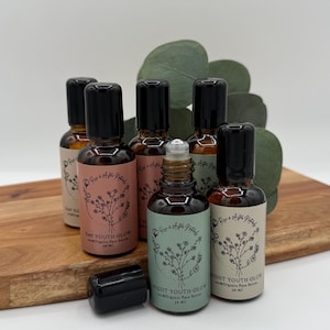 May include: A collection of small, brown glass bottles with black caps, labeled with text and floral designs. The bottles are arranged on a wooden cutting board with green foliage in the background. The labels read "Day Youth Glow" and "Night Youth Glow".