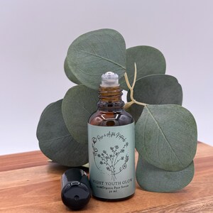 May include: A brown glass bottle with a rollerball applicator and a black cap, labeled "NIGHT YOUTH GLOW 100% Organic Face Serum 10 ml." The bottle sits on a wooden surface with green leaves in the background.