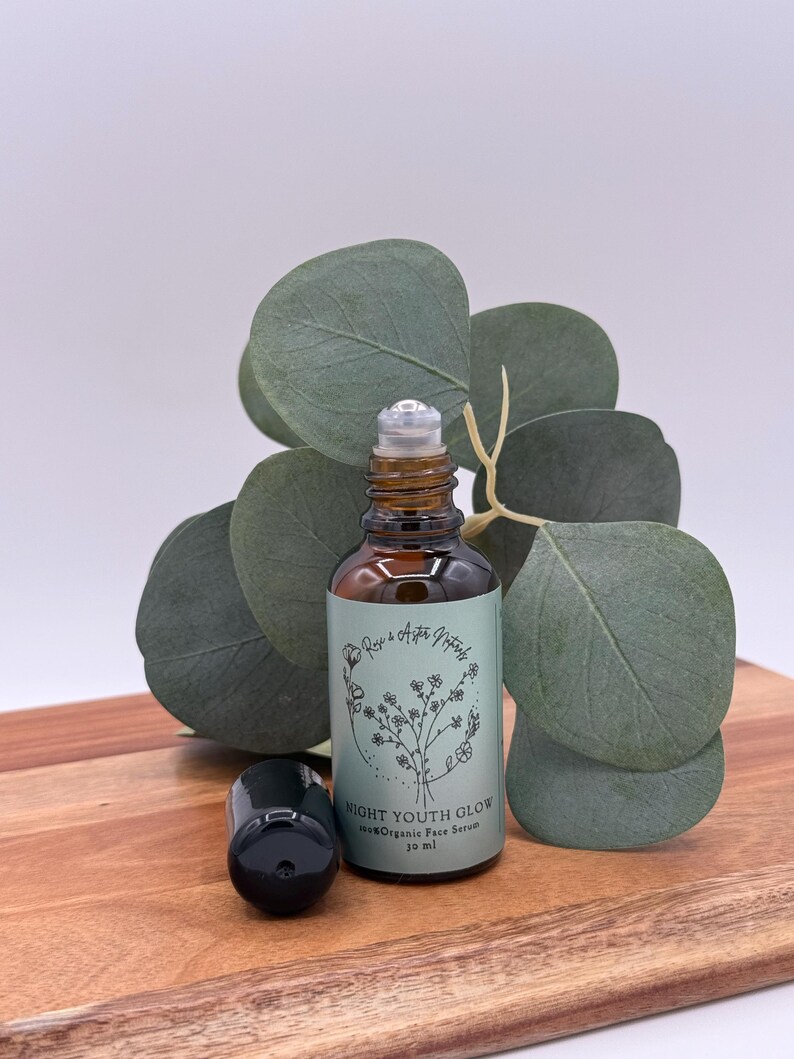 May include: A brown glass bottle with a rollerball applicator and a black cap, labeled "NIGHT YOUTH GLOW 100% Organic Face Serum 10 ml." The bottle sits on a wooden surface with green leaves in the background.