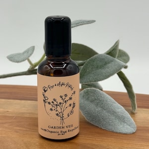 May include: A brown glass bottle with a black cap, labeled "Garden Veil 100% Organic Bug Repellent." The label features floral illustrations and the brand name "Rose & Aster Naturals." The bottle contains 30 ml of product and is set against a wooden surface.