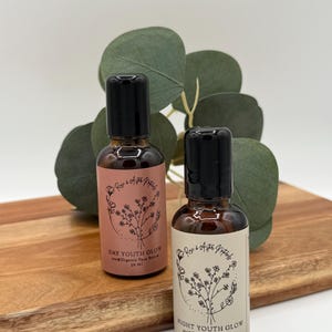 May include: Two small brown glass bottles with black caps, labeled "DAY YOUTH GLOW" and "NIGHT YOUTH GLOW" with floral illustrations. The bottles are on a wooden cutting board with green foliage in the background. Each bottle contains 30 ml of organic face serum.