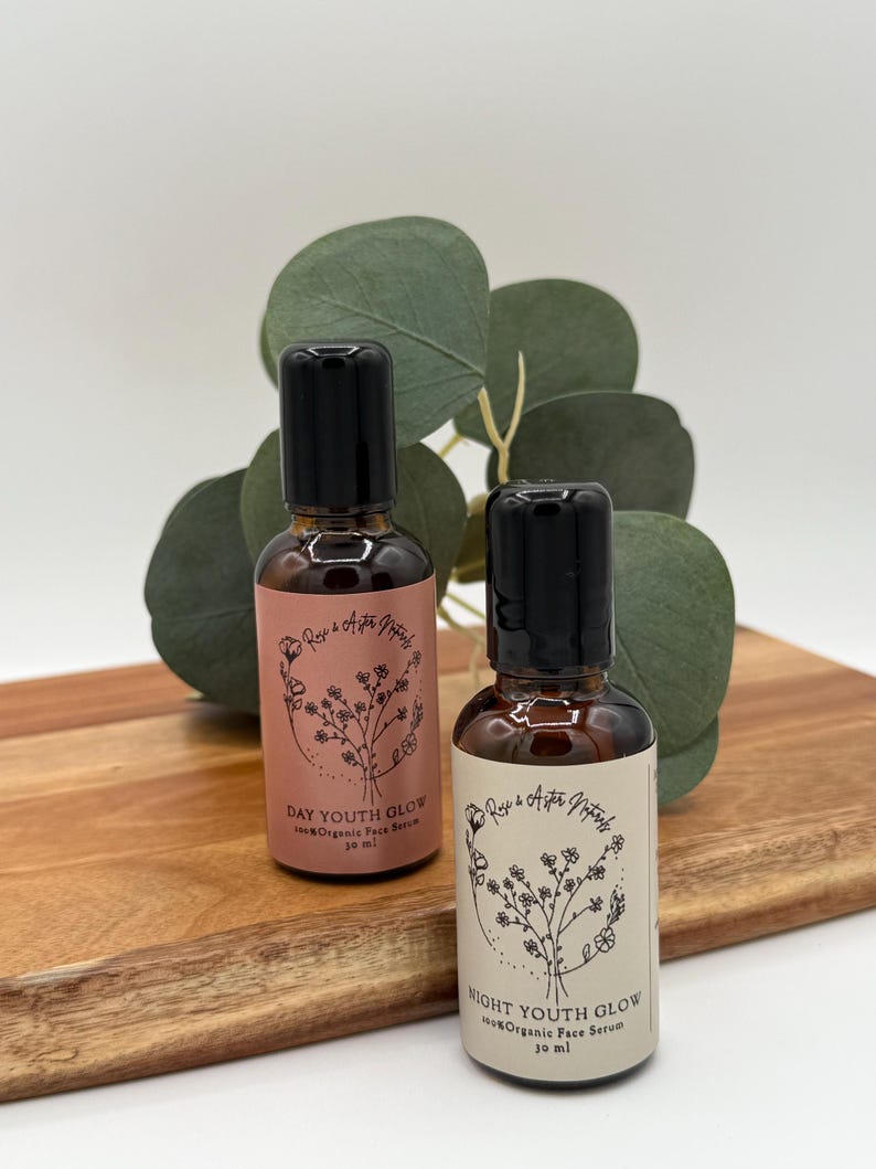 May include: Two small brown glass bottles with black caps, labeled "DAY YOUTH GLOW" and "NIGHT YOUTH GLOW" with floral illustrations. The bottles are on a wooden cutting board with green foliage in the background. Each bottle contains 30 ml of organic face serum.