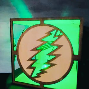 May include: A wooden box with a green interior and a circular design featuring a lightning bolt. The lightning bolt is tan, and the box has a square frame. The box is illuminated with green light.