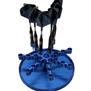 May include: A blue circular dart stand with multiple radial arms featuring circular cutouts for storage. Five black and blue darts with patterned flights are inserted into the central section of the stand, which has a smooth, matte finish.