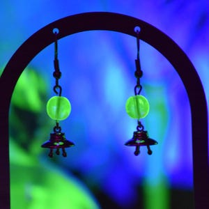 May include: A pair of unique earrings featuring a neon green bead and a small, dark-colored UFO charm. The earrings hang from a black metal display stand against a blurred blue and green background. The earrings are designed to catch the eye.