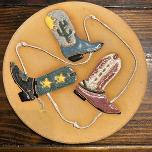 Könnte beinhalten: Ein rundes, hellbraunes Dekorationsobjekt mit drei Cowboy-Stiefeln in Blau, Grün und Rot. Die Stiefel sind durch ein weißes Seil verbunden und haben Kaktus-, Stern- und dekorative Designs. Der Hintergrund ist einfarbig hellbraun.