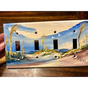Könnte beinhalten: Handbemalte Lichtschalterabdeckung mit einer Küstenlandschaft. Das Kunstwerk zeigt einen Strand mit blauem Wasser, Sanddünen und hohem Gras. Zwei weiße Vögel fliegen am Himmel. Die Abdeckung hat vier Schalteröffnungen und Schraubenlöcher.