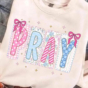 Könnte beinhalten: Ein cremefarbenes Sweatshirt mit dem Wort PRAY in großen, dekorativen Buchstaben. Jeder Buchstabe hat ein einzigartiges Muster, darunter rosa Herzen, blaue Blumen, rosa Streifen und blaue Karos, akzentuiert mit rosa Schleifen und kleinen Sternen.