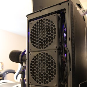 May include: A black computer case with three large, circular fans with a honeycomb pattern. The fans are stacked vertically. A microphone is visible to the left. The case is on a wooden surface.