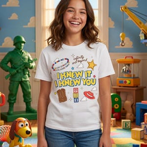May include: A white short-sleeve t-shirt featuring a colorful graphic print with the text "I KNEW IT I KNEW YOU" and "infinity and beyond," surrounded by illustrations of a beaded bracelet, a gold star, a brown boot, alphabet blocks, and a red hat.