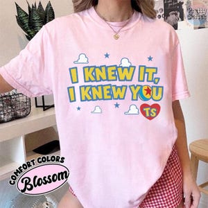 May include: A light pink short-sleeve t-shirt featuring the yellow and blue text I KNEW IT, I KNEW YOU, a red heart with the letters TS, and small blue stars and white cloud graphics printed on the front center of the garment.