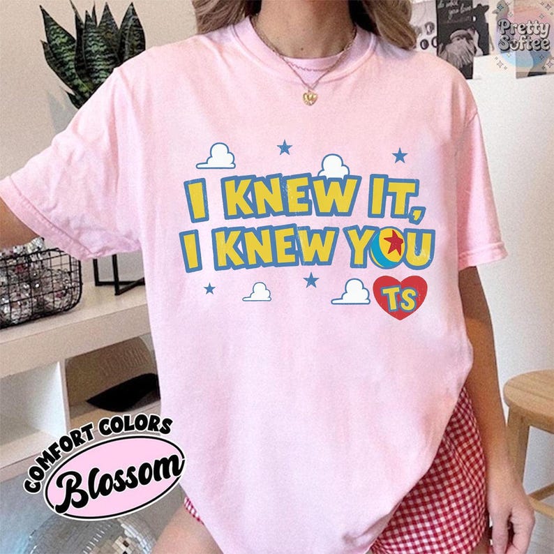 May include: A light pink short-sleeve t-shirt featuring the yellow and blue text I KNEW IT, I KNEW YOU, a red heart with the letters TS, and small blue stars and white cloud graphics printed on the front center of the garment.