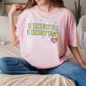 May include: A light pink short-sleeved t-shirt featuring the yellow text I KNEW IT, I KNEW YOU, a red heart with the letters TS, and a small ball graphic. The design is surrounded by small blue stars and white cloud outlines on the front chest.