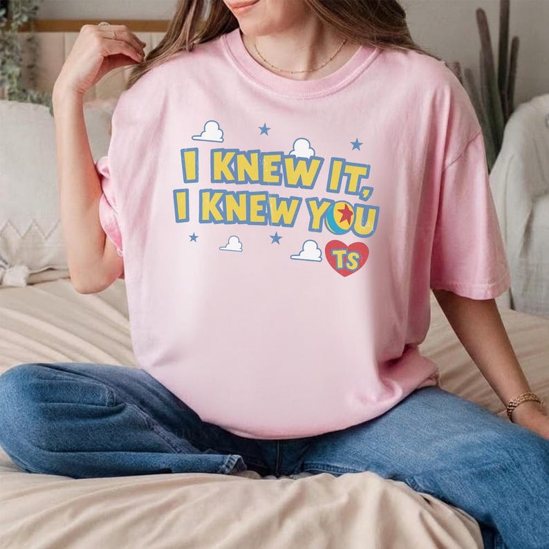 May include: A light pink short-sleeved t-shirt featuring the yellow text I KNEW IT, I KNEW YOU, a red heart with the letters TS, and a small ball graphic. The design is surrounded by small blue stars and white cloud outlines on the front chest.
