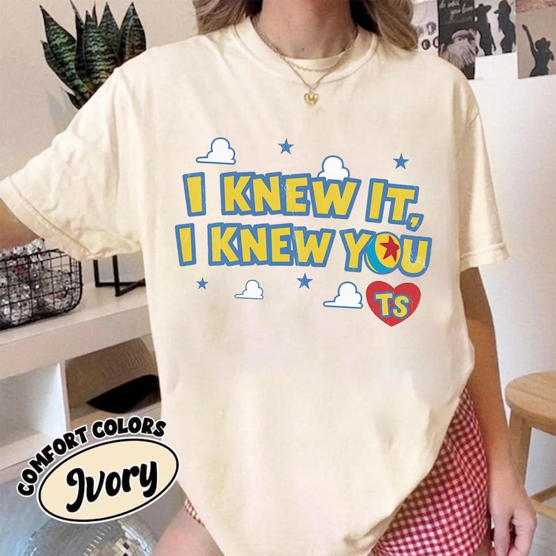 May include: An ivory short-sleeve t-shirt featuring the blue text I KNEW IT, I KNEW YOU with a red heart containing the yellow letters TS. The design includes small blue stars and white cloud outlines, with a ball graphic replacing the letter O.