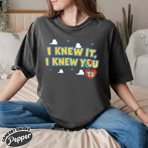 May include: A charcoal gray short-sleeve t-shirt featuring the yellow text I KNEW IT, I KNEW YOU with a colorful ball graphic, small white clouds, blue stars, and a red heart containing the letters TS printed on the front chest area.