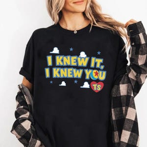 May include: A black short-sleeved t-shirt featuring the yellow text I KNEW IT, I KNEW YOU with a blue and red ball icon replacing the letter O. The design includes white cloud shapes, blue stars, and a red heart with the yellow letters TS.