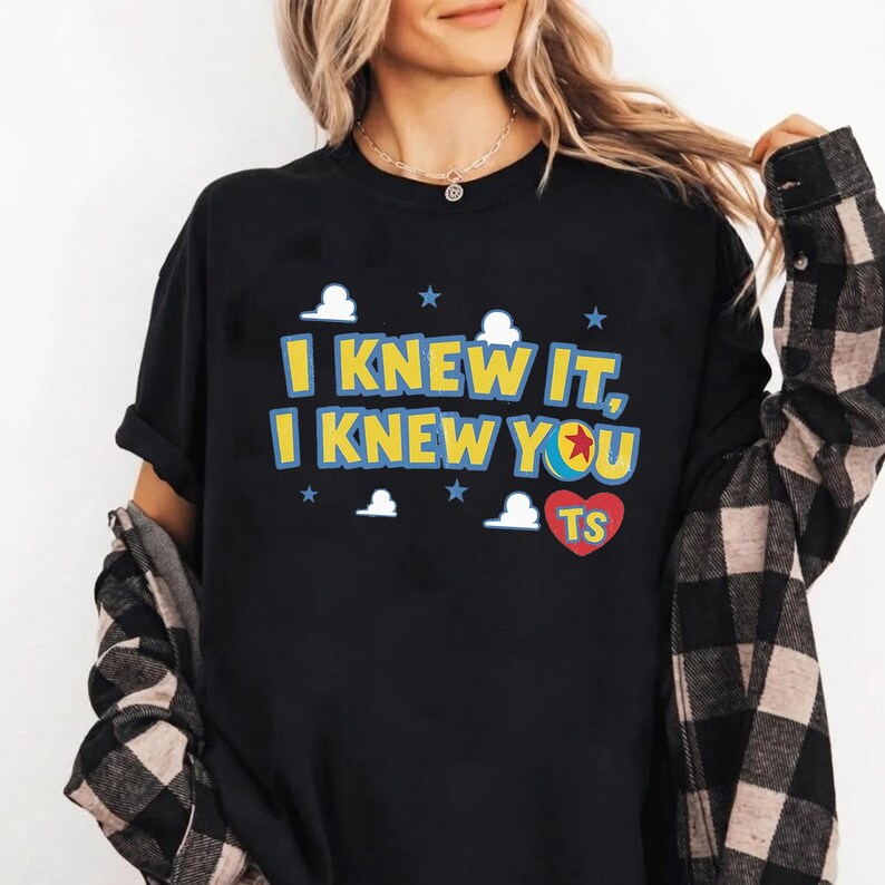 May include: A black short-sleeved t-shirt featuring the yellow text I KNEW IT, I KNEW YOU with a blue and red ball icon replacing the letter O. The design includes white cloud shapes, blue stars, and a red heart with the yellow letters TS.