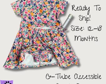 May include: A floral print onesie dress with short sleeves and a G-tube accessible opening. The fabric features a colorful pattern of pink, orange, and purple flowers. Text on the image reads: Ready To Ship! Size: 12-18 Months, G-Tube Accessible Onesie Dress, Love, Wilson.