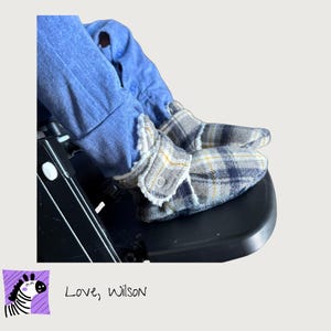 May include: Soft baby booties featuring a grey, navy blue, and yellow plaid pattern. The booties have a white faux-shearling lining and a side snap button closure. They are worn over blue fabric pants, showcasing the textured fabric construction.