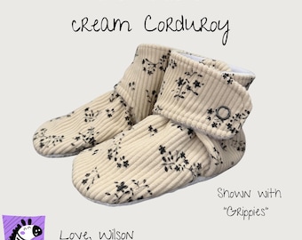 May include: A pair of cream corduroy booties featuring a black vine floral pattern. The text on the image reads: Vine floral on cream Corduroy, Shown with Grippies, Love, Wilson. The booties have a fold-over cuff secured with a small metal snap.