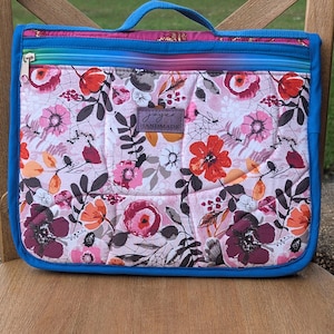 May include: Quilted floral print bag with a blue handle and trim. The bag features a colorful floral pattern with pink, red, and orange flowers on a light pink background. The bag has a zipper closure and a small tag that says "handmade".
