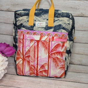 May include: A handmade tote bag with a navy blue background featuring a white bird pattern. The front pocket displays a pink and red palm tree design. It has yellow handles and the text "YOU ARE BEAUTIFUL".