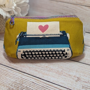 May include: Mustard yellow zippered pouch featuring a typewriter design with a pink heart on the paper. The typewriter is blue and black. The zipper pull is a rainbow color. The pouch is on a wooden surface.
